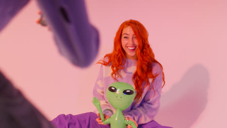 Studio-Shot-Of-Woman-Taking-Photo-Of-Friend-Playing-With-Toy-Inflatable-Alien-On-Mobile-Phone-Against-Pink-Background